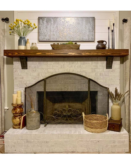 Rustic wood mantel with dark metal brackets mounted on white brick fireplace featuring farmhouse style decor and accessories