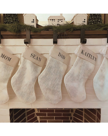 Rustic wooden mantel with black metal stocking holders displaying cream burlap Christmas stockings personalized with famil...