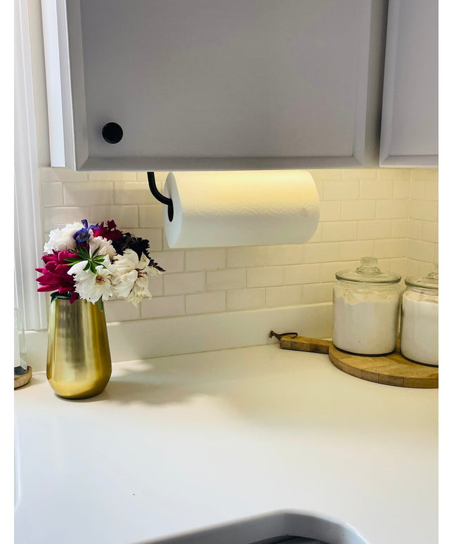 Black metal Hyde Hall Paper Towel Holder mounted under white kitchen cabinet with white paper towel roll on marble countertop