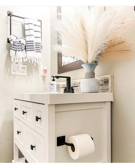 Black metal toilet paper holder mounted on white bathroom vanity cabinet with farmhouse style drawers and black hardware