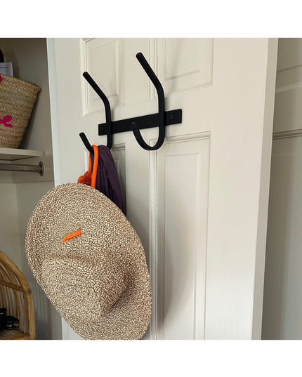 Black metal Meredith Petite Double J Coat Rack by Industrial Farm Co mounted on white door with woven hat and clothing