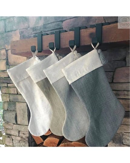 Four neutral-toned burlap Christmas stockings hanging from black metal Annie Stocking Holders on rustic brick fireplace ma...