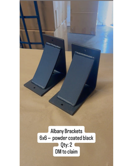 Two black powder coated Albany 6x6 metal brackets from Industrial Farm Co displayed on wooden surface in warehouse setting