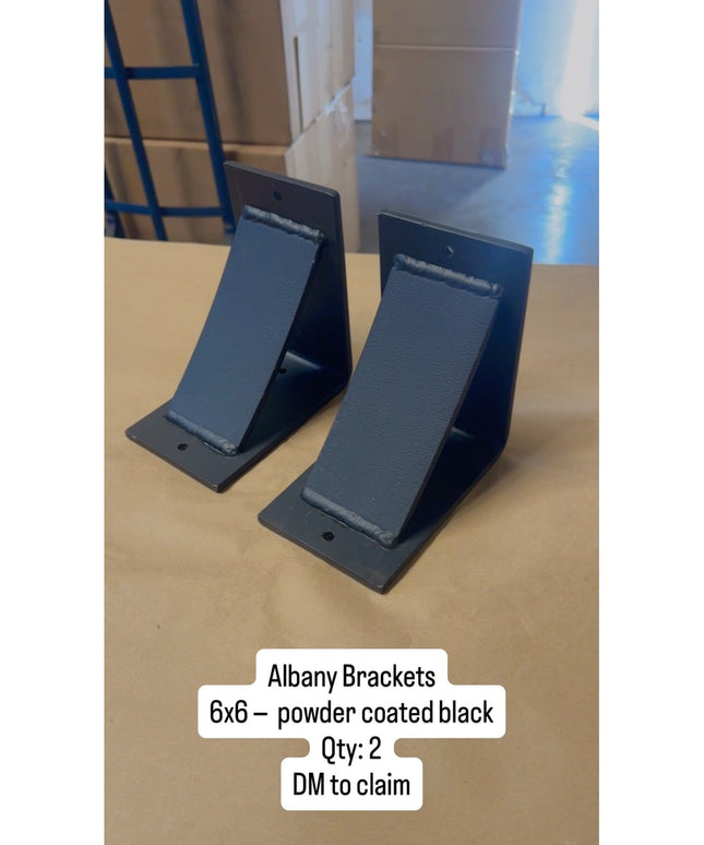 Two black powder coated Albany 6x6 metal brackets from Industrial Farm Co displayed on wooden surface in warehouse setting