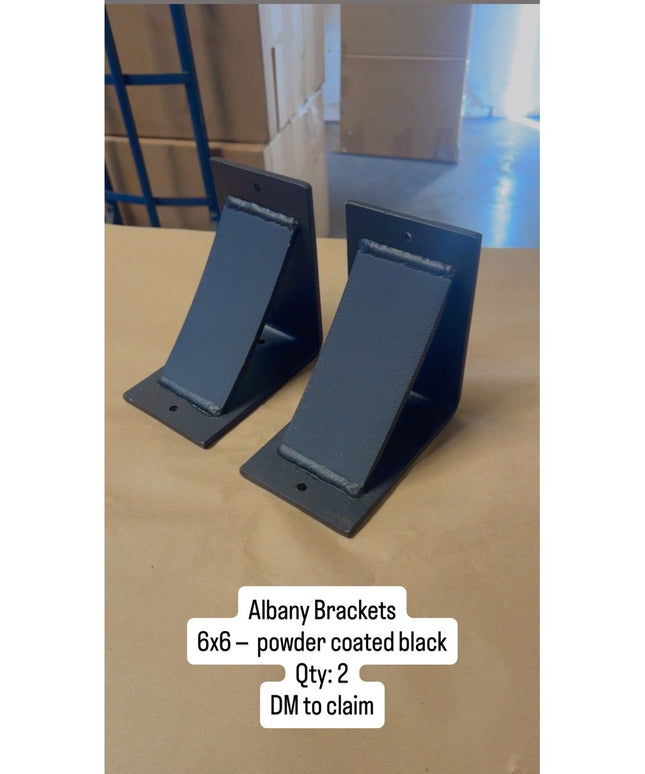 Two black powder coated Albany 6x6 metal brackets from Industrial Farm Co displayed on wooden surface in warehouse setting