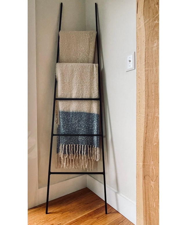 Black metal tapered farmhouse ladder by Industrial Farm Co displaying cream and blue textured blankets in corner against w...