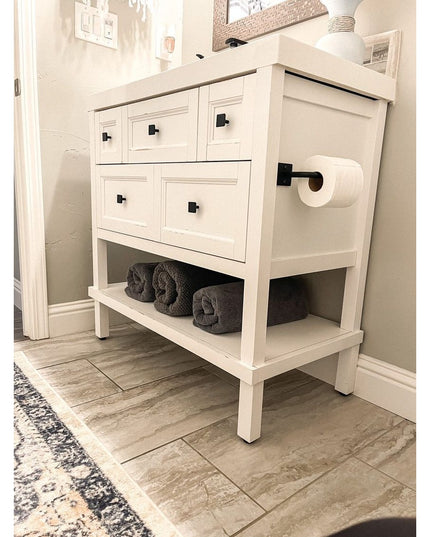 White farmhouse bathroom vanity with five drawers, black hardware, open bottom shelf with gray towels, and black toilet pa...