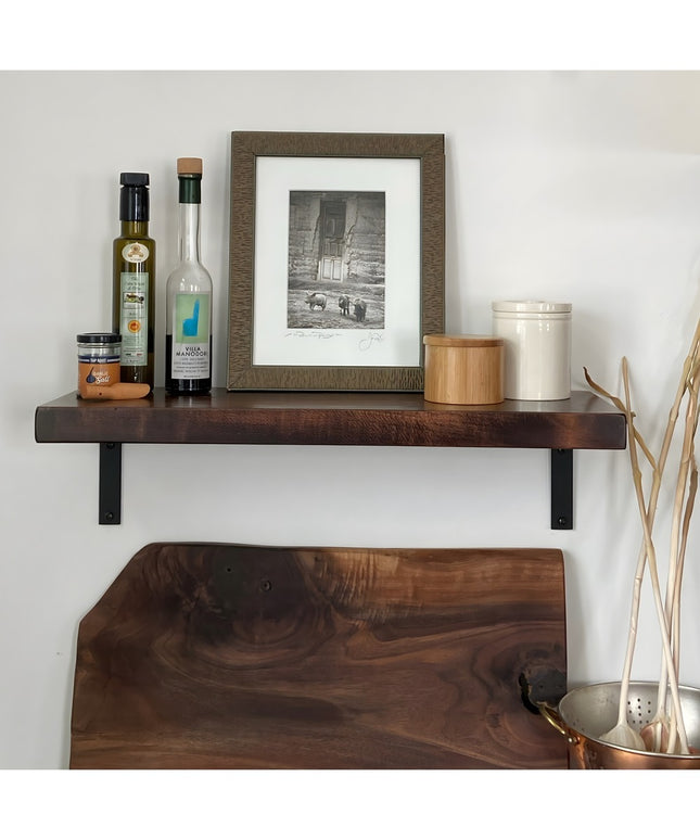 Dark walnut floating shelf with black steel L-brackets displaying kitchen items and framed artwork on white wall