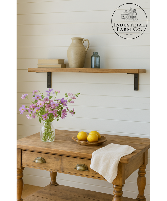 Wooden table with a vase of flowers, lemons, and a towel, with a shelf above featuring a pitcher and bottles against a white paneled wall.
