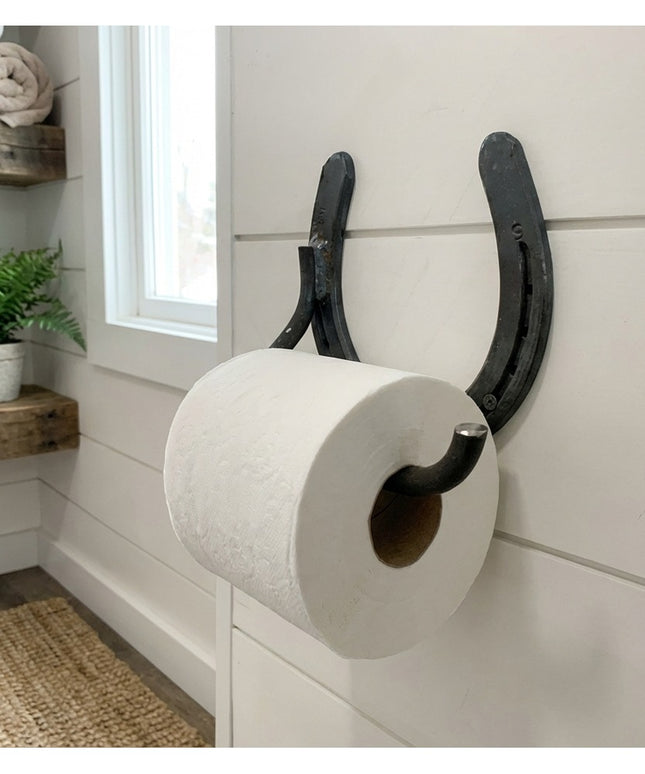 Rustic black horseshoe toilet paper holder mounted on white shiplap bathroom wall with farmhouse decor and floating wood s...